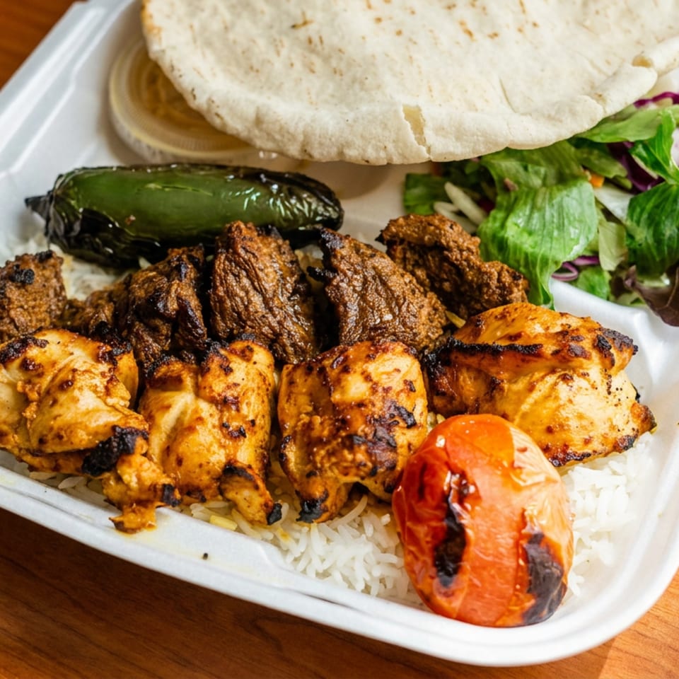 Grilled chicken and beef kebabs with charred vegetables, rice, salad, and flatbread in a takeout tray.