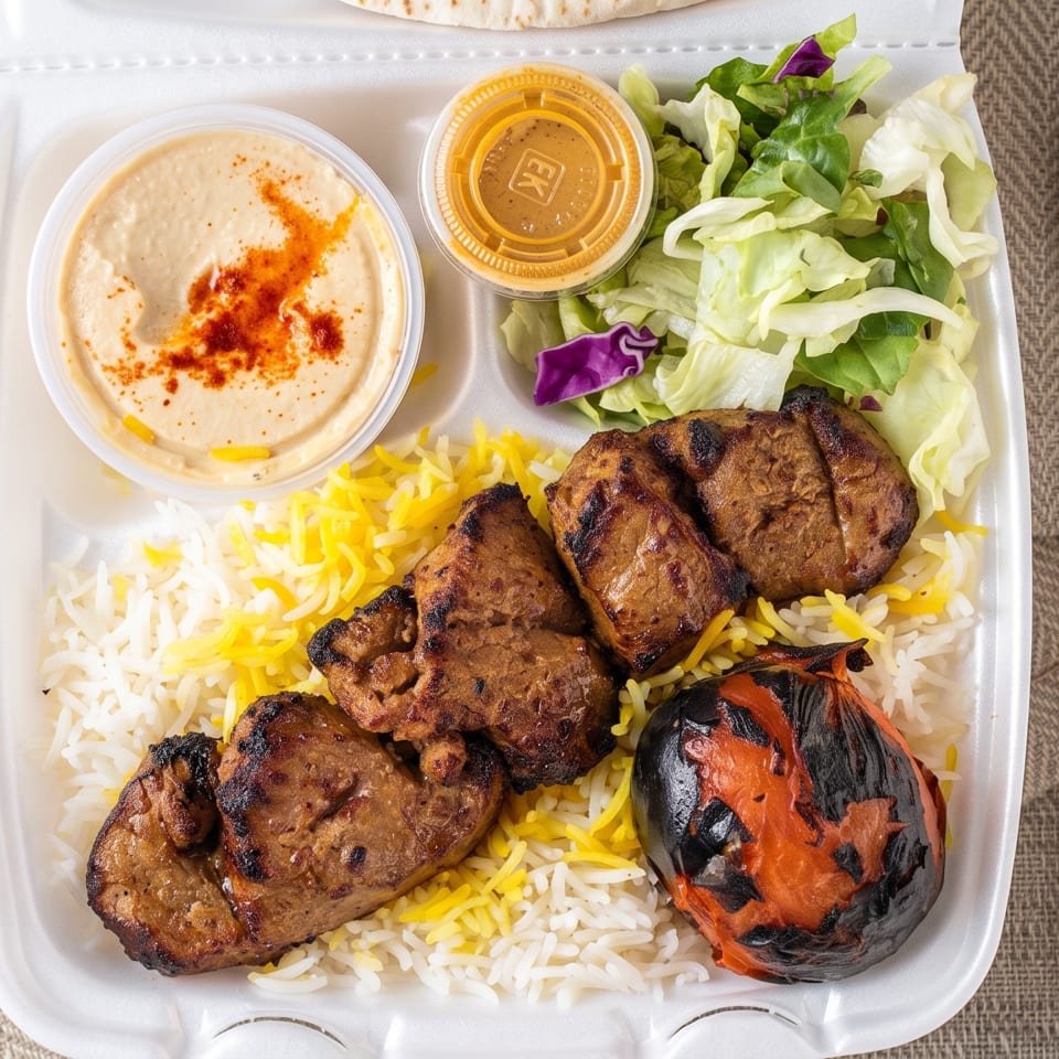 Grilled meat and charred eggplant served over saffron rice with hummus, salad, and dressing in a takeout container.