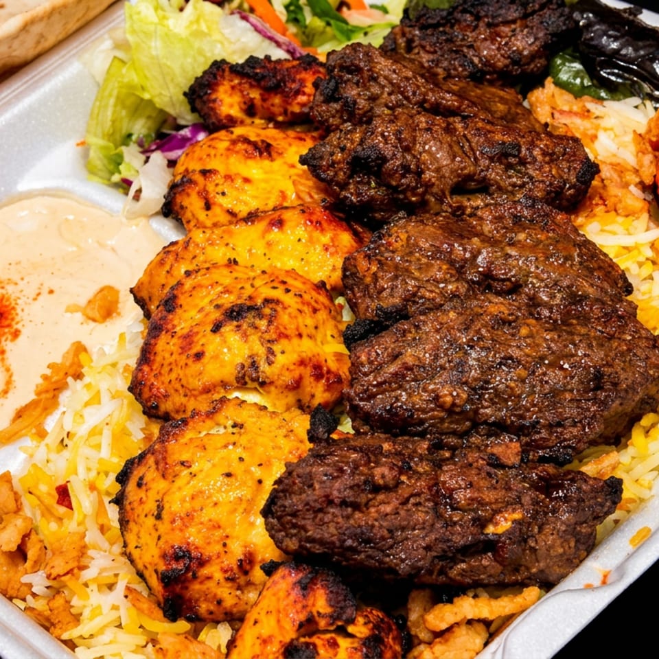 Mixed grill platter with grilled kebabs, chicken, rice, salad, flatbread, hummus, and roasted tomato in a takeaway tray.